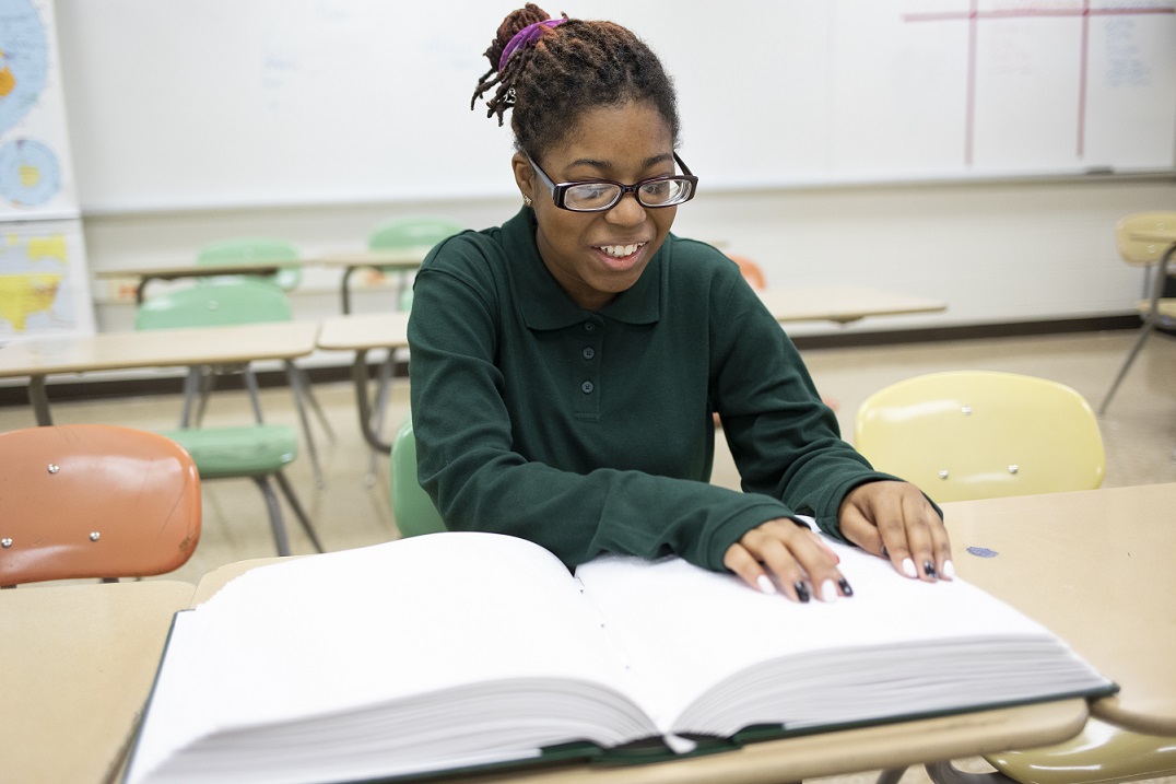 A girl reads from a Braille book; she is smiling.